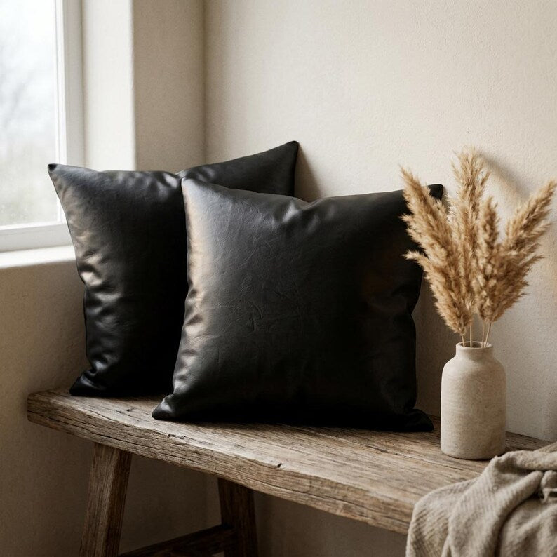 Two black vegan leather pillow covers on a rustic wooden bench next to a window, with a textured vase and dried pampas grass.