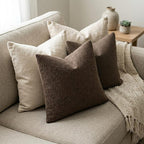 Two brown boucle pillow covers styled with two off-white cushions on a light beige sofa, featuring a knitted throw blanket.
