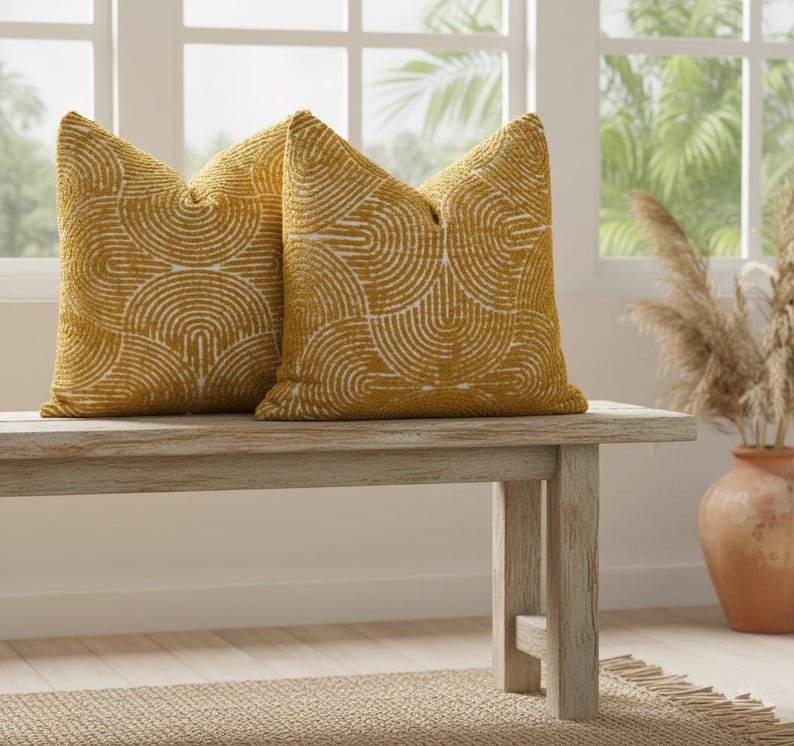 Two yellow patterned pillows on a wooden bench with a vase and plants in the background.