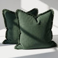 Set of 2 olive green linen throw pillow covers with fringe trim on all four sides, styled on a white surface