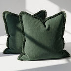 Set of 2 olive green linen throw pillow covers with fringe trim on all four sides, styled on a white surface