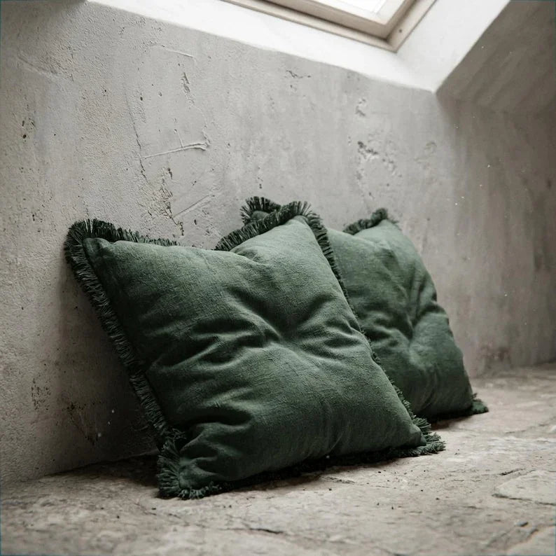 Two olive green linen fringe pillow covers leaning against a textured concrete wall in a natural light setting