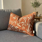 Burnt orange botanical chenille lumbar pillow cover with trailing leaf print, styled on gray sofa with olive tree decor