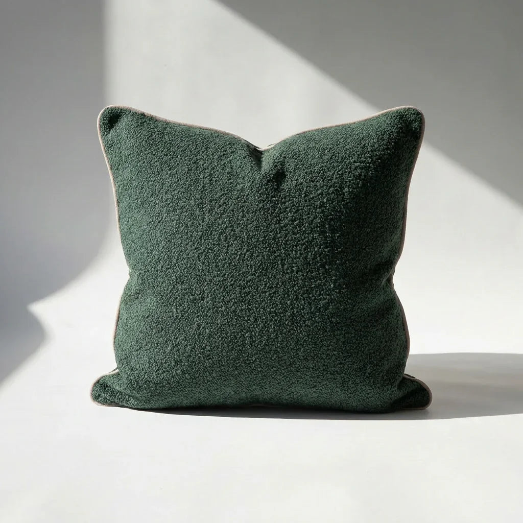 Dark green square boucle pillow cover with beige piping, textured fabric on white surface with sunlight shadows.
